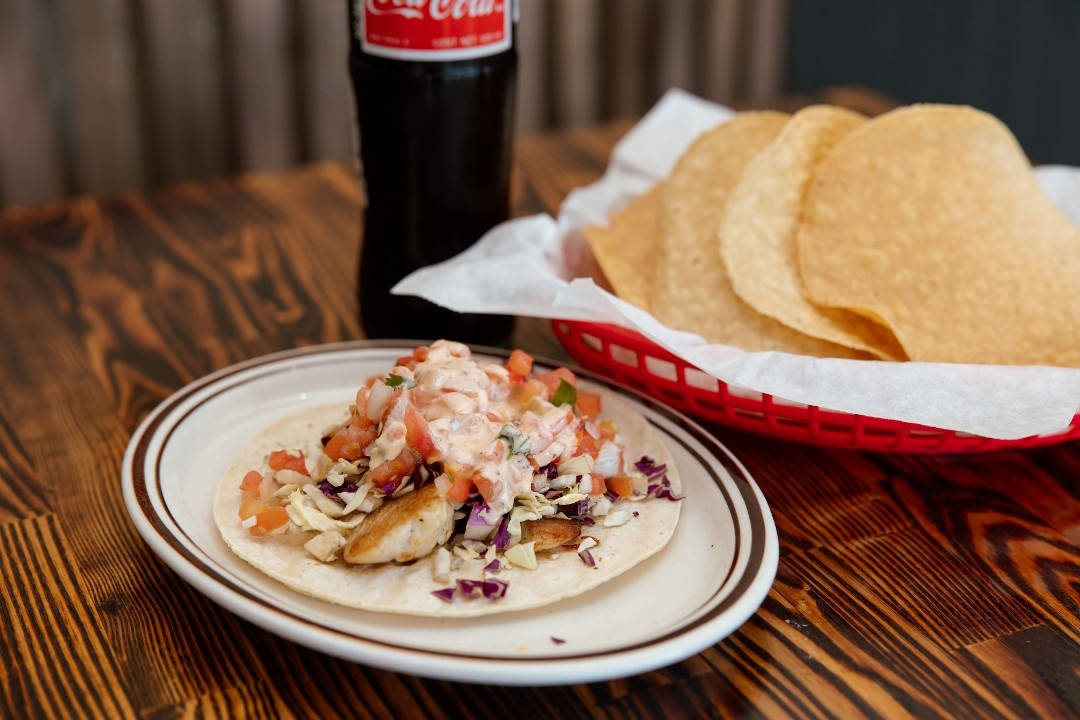 A fish taco on a soft tortilla with shredded cabbage and pico de gallo, on a rimmed plate beside a basket of chips and a bottle of Coca-Cola.