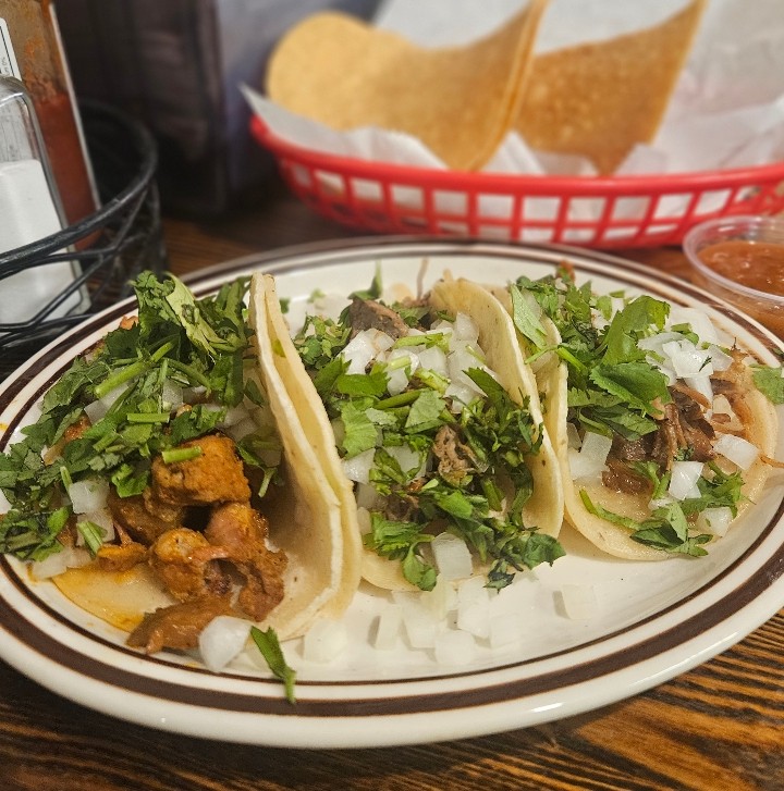 Three small street tacos on soft corn tortillas topped with chopped onion and cilantro, on a diner plate beside a basket of tortilla chips.