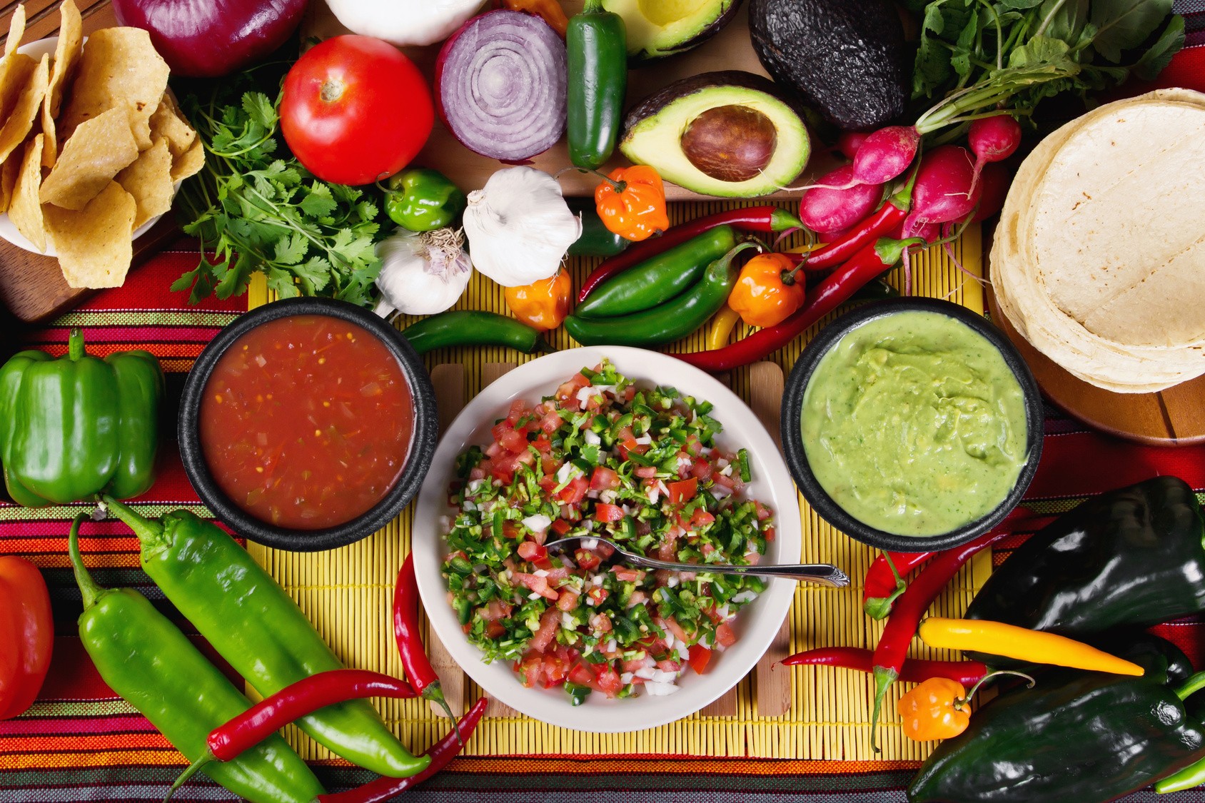 Fresh chiles, tomatoes, onion, avocado and tortillas laid out around bowls of red salsa, green salsa and pico de gallo.
