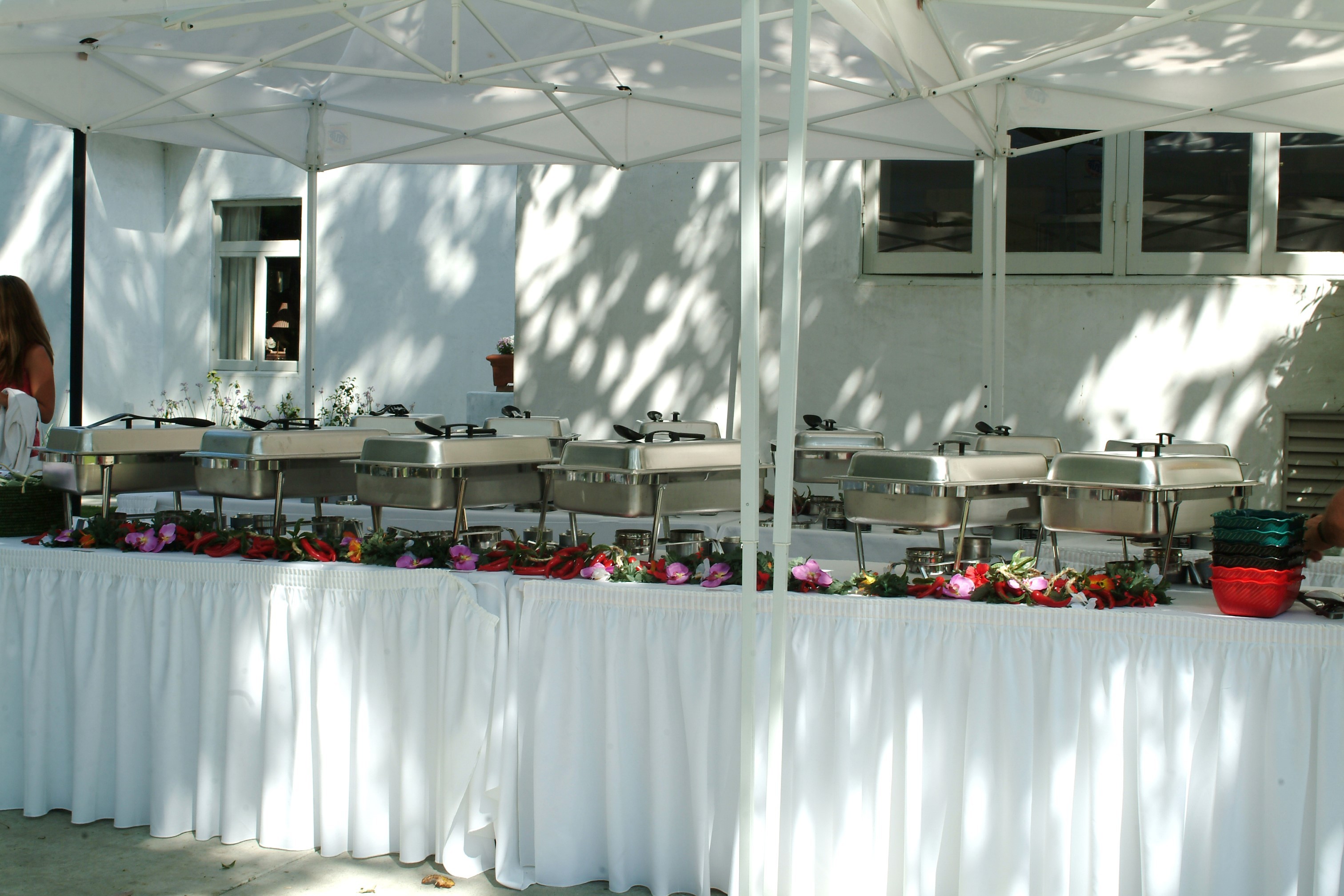 A long white-draped buffet under a marquee, chafing dishes lined up and trimmed with flowers, ready for guests.