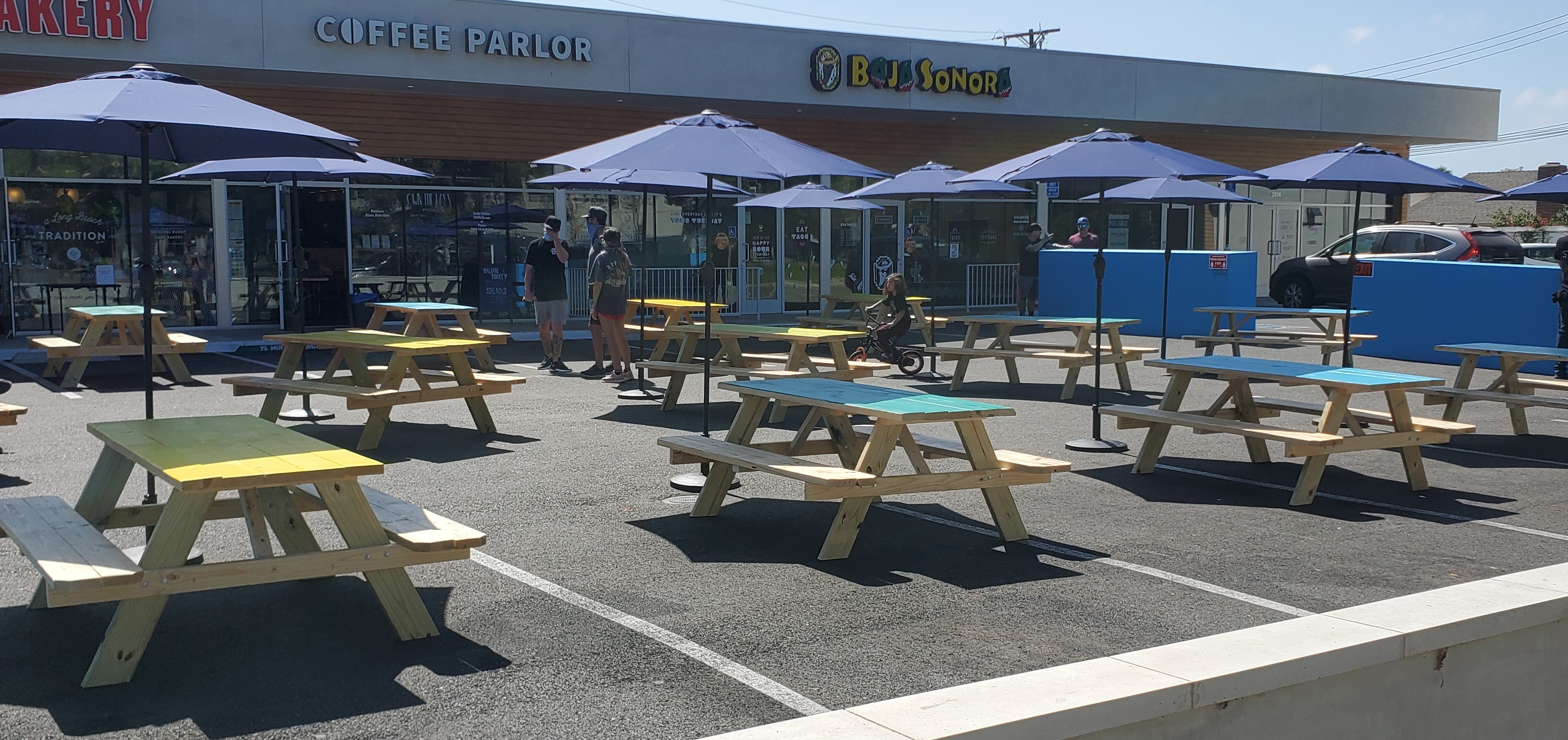 The restaurant's patio and forecourt: rows of picnic tables under blue umbrellas in front of the storefront on a clear day.
