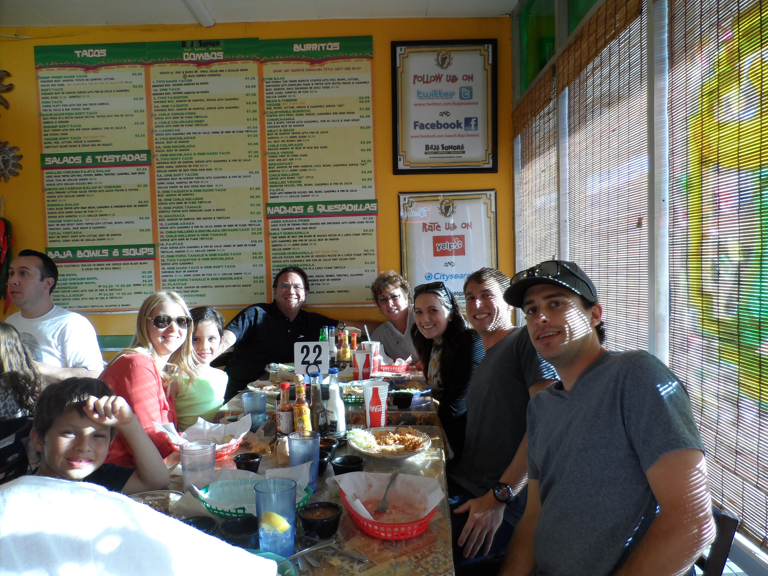 A group of guests smiling around a table covered in food and drinks, beneath the restaurant's hand-painted menu board.