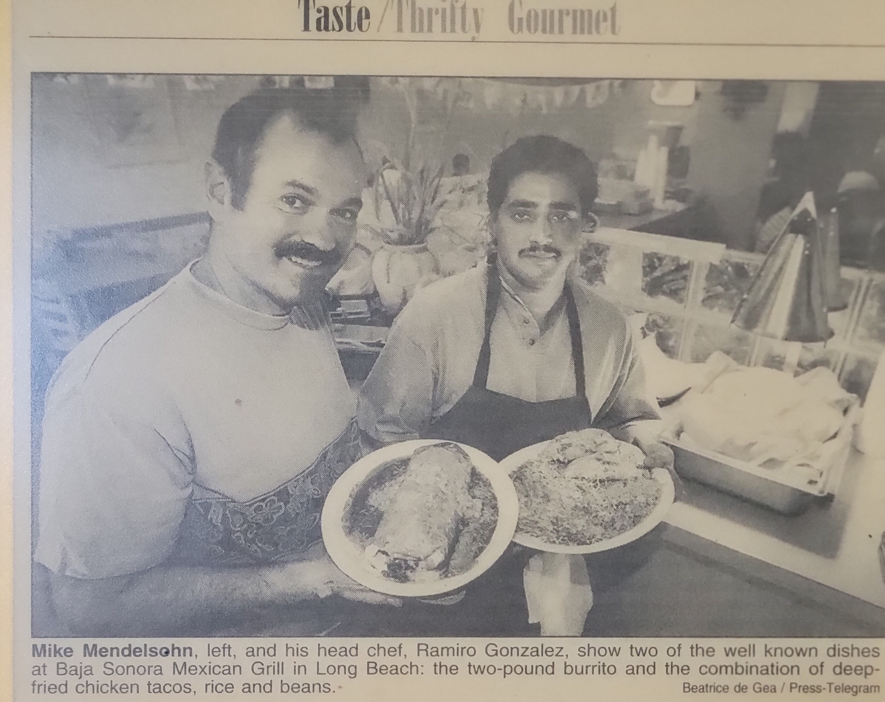 A newspaper clipping headlined Taste / Thrifty Gourmet, showing Mike Mendelsohn and head chef Ramiro Gonzalez each holding a plate — one a large burrito, one deep-fried tacos with rice and beans.