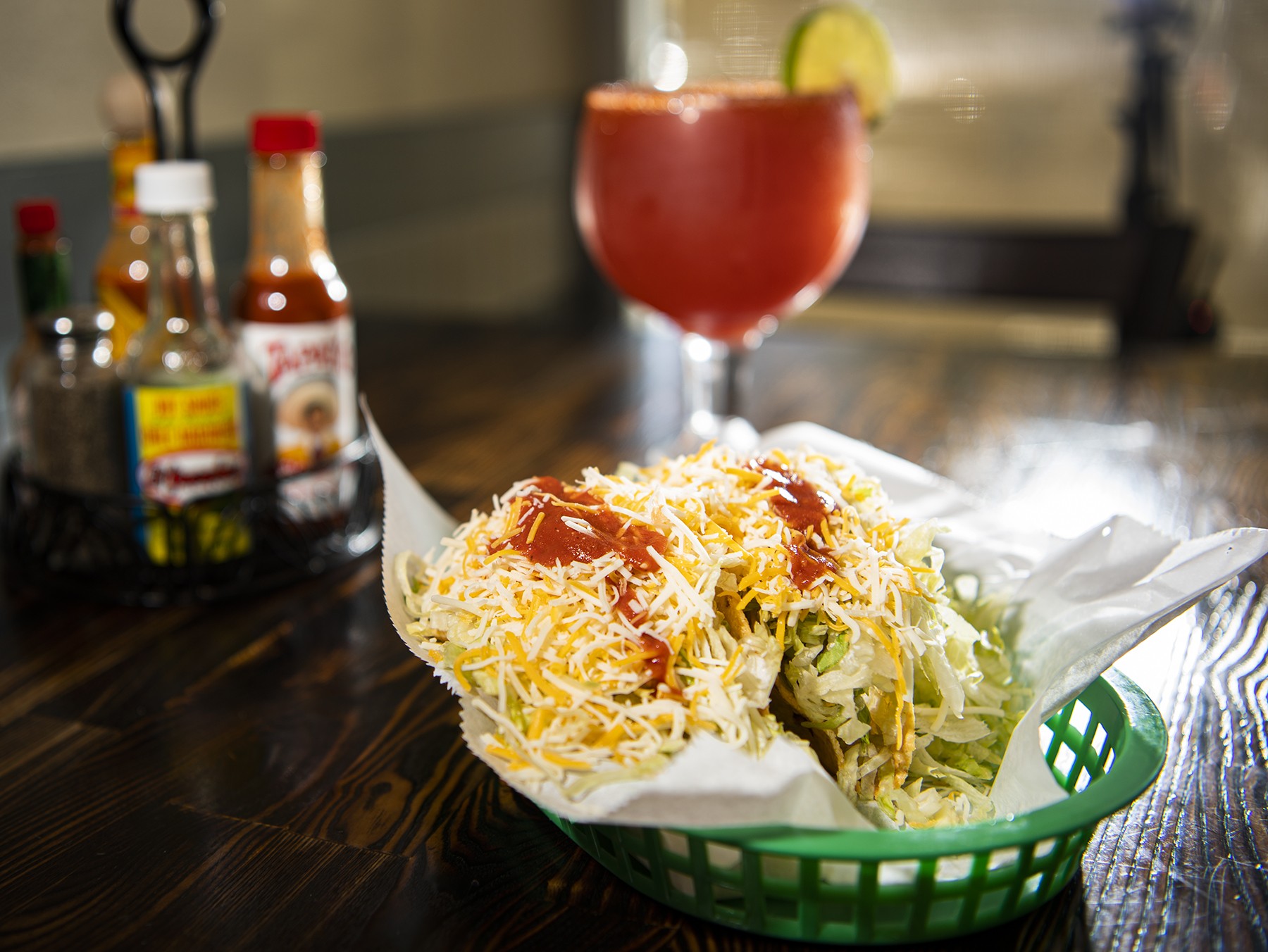 A tostada piled with lettuce, cheese and salsa in a green basket on a dark wood table, with hot sauce bottles and a tall michelada beside it.