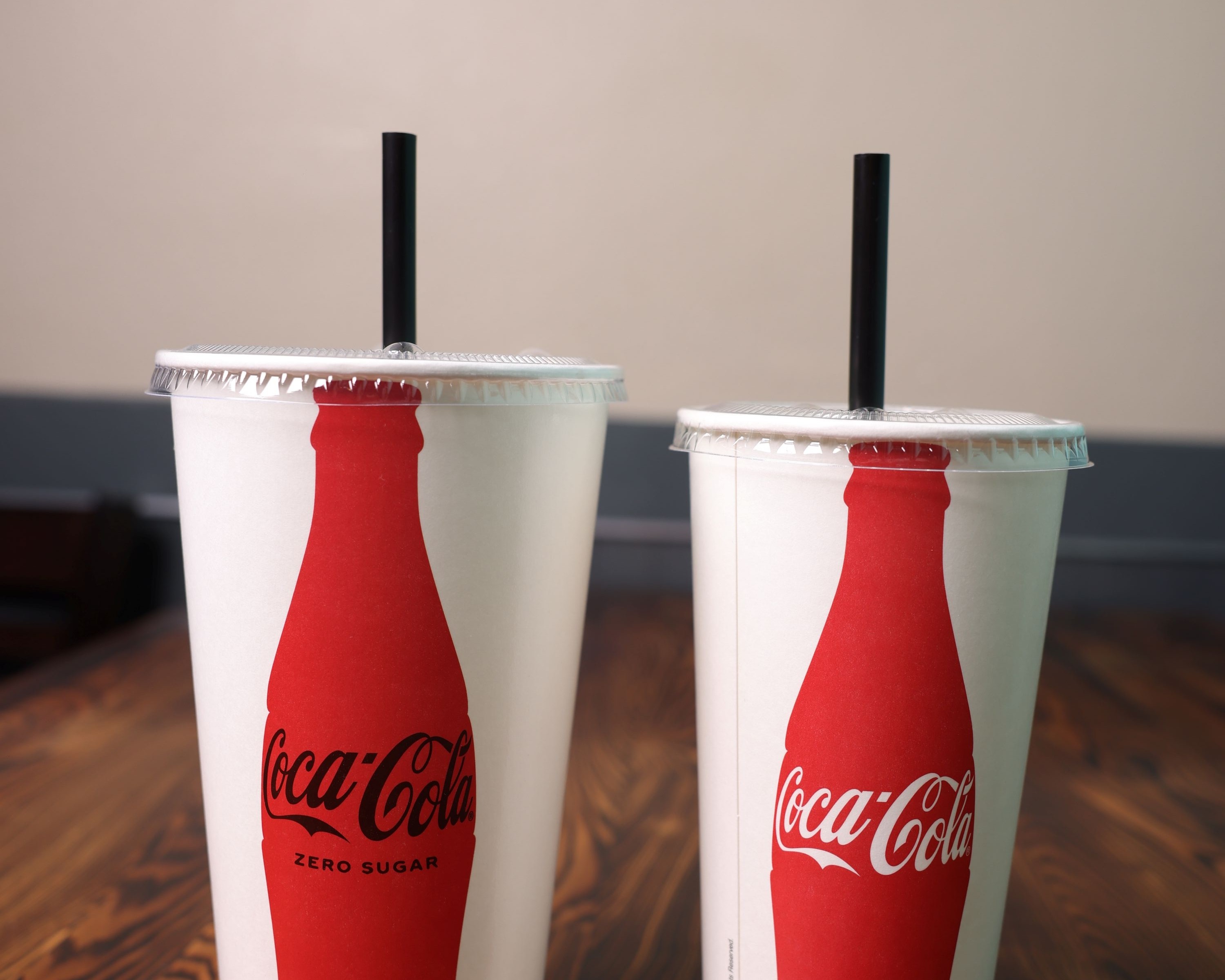 Two tall Coca-Cola cups with straws standing on a polished dark wood table in the dining room.