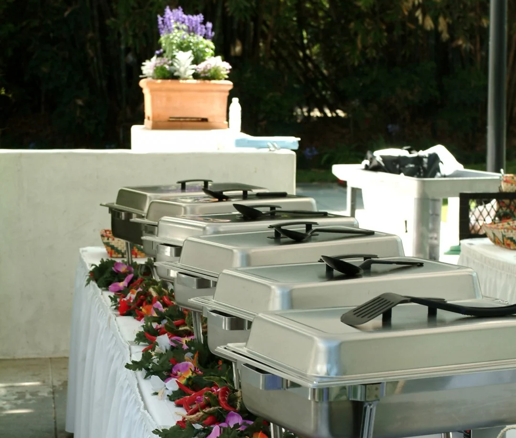 A row of stainless chafing dishes set up outdoors along a white buffet counter trimmed with flowers.